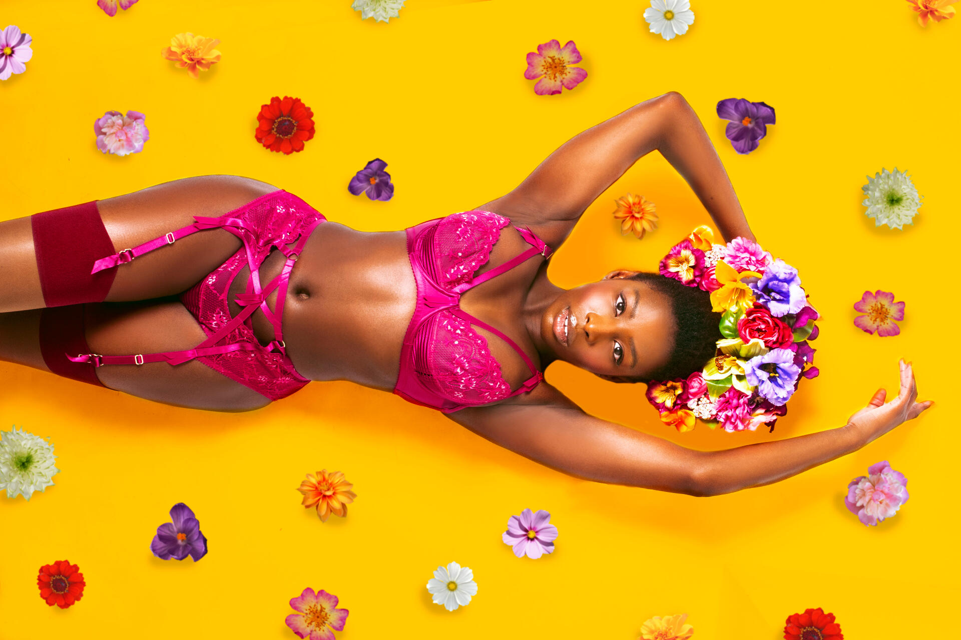 Red Tongued Raven wearing lingerie by Dita Von Teese, lying against a yellow background surrounded by flowers